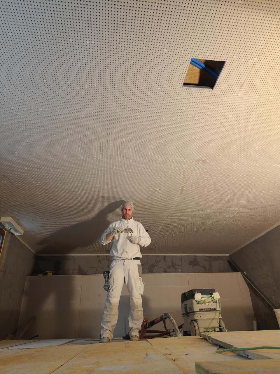 Eine Person in weisser Arbeitskleidung steht in einem unfertigen Raum mit perforierter Decke neben einem Staubsauger.
