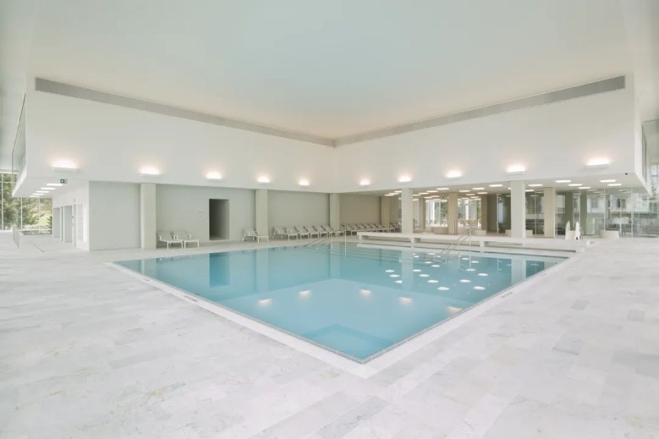 Ein Hallenbad, umgeben von einer grossen, weiss gefliesten Terrasse unter heller, minimalistischer Architekturbeleuchtung.