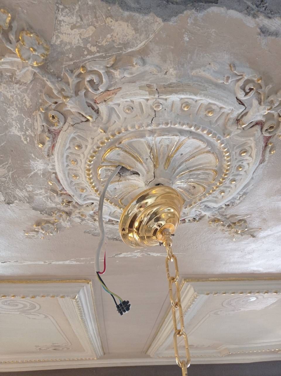 Ein goldener Kronleuchterbaldachin mit Kette hängt von einem reich verzierten, patinierten Deckenrosette.
