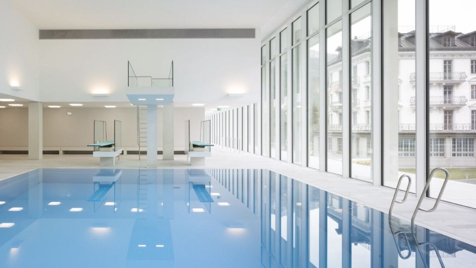 Modernes Hallenbad mit blauem Wasser, Sprungtürmen und raumhohen Fenstern mit Blick auf ein Gebäude.