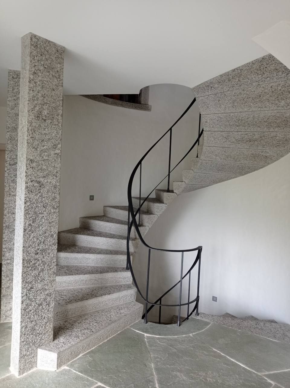 Eine spiralförmige Granittreppe mit dunklem Metallgeländer vor weissen Wänden und grauem Steinboden.