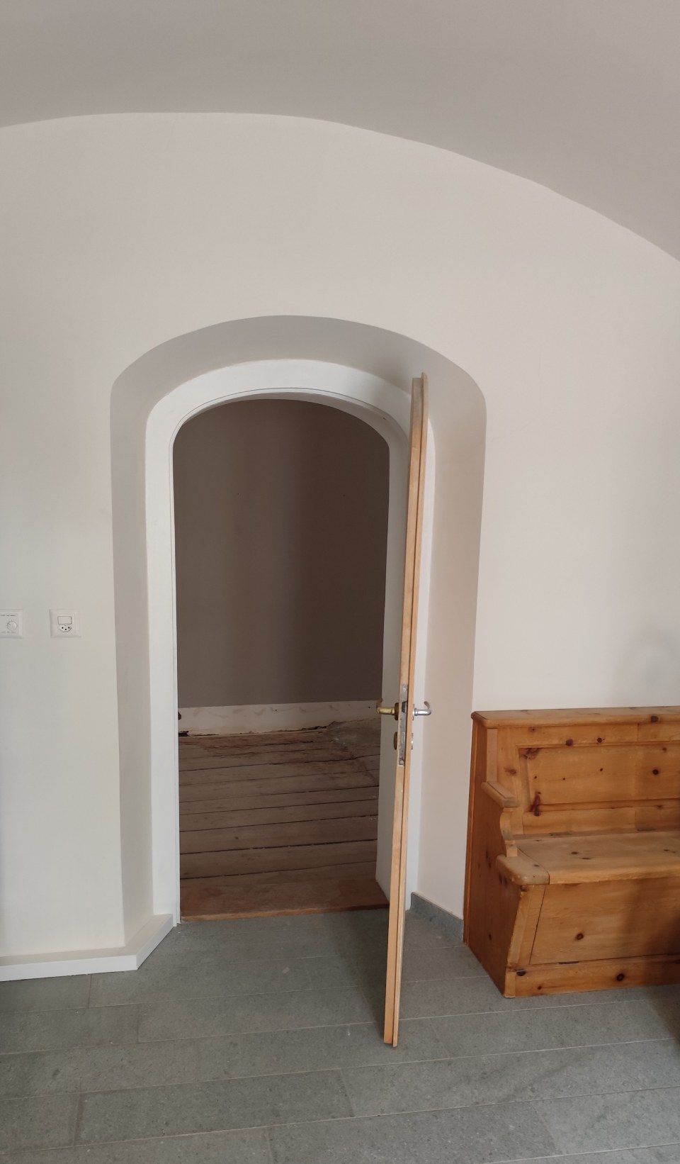 Durch einen Bogengang gelangt man in einen Raum mit Holzboden, neben dem sich eine Holzbank auf einem Steinboden befindet.