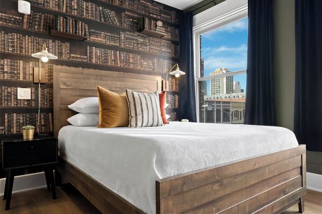 Cozy bedroom with bookshelf wallpaper, white bed, and city view through a large window