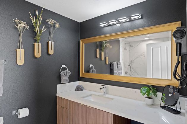 Modern bathroom vanity with wood cabinets, white countertop, gold-framed mirror, and wall decor