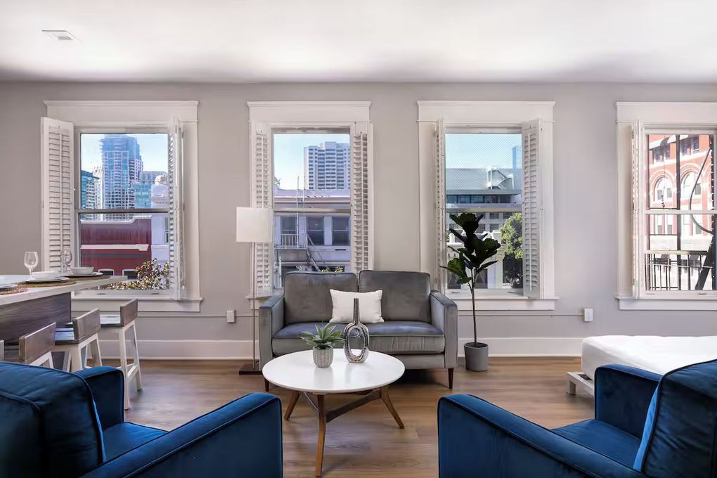 Modern living room with blue chairs, a gray sofa, and large windows overlooking city buildings.