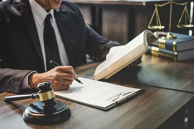 Un abogado con traje firmando un documento, con un mazo, libros de leyes y una balanza de justicia sobre un escritorio.