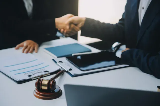 Dos personas de traje se dan la mano en una mesa con documentos legales, un mazo y una tableta.