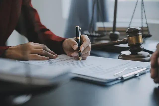 Una persona firma documentos legales con un bolígrafo en un escritorio, con una balanza de la justicia y un mazo al fondo.