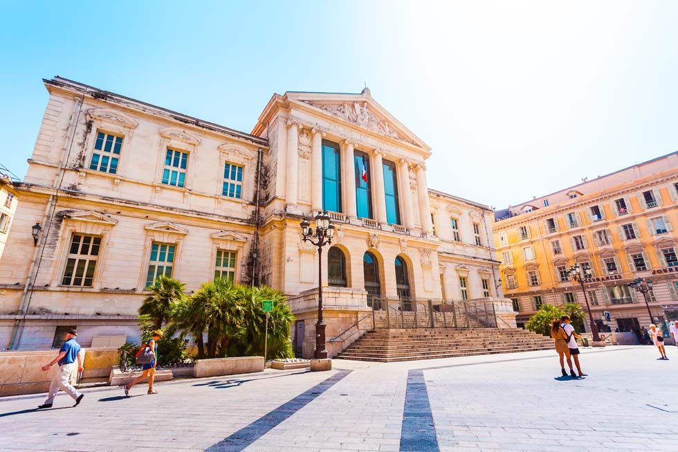 Bâtiment historique et place baignés de soleil, avec quelques personnes qui se promènent sous un ciel bleu éclatant