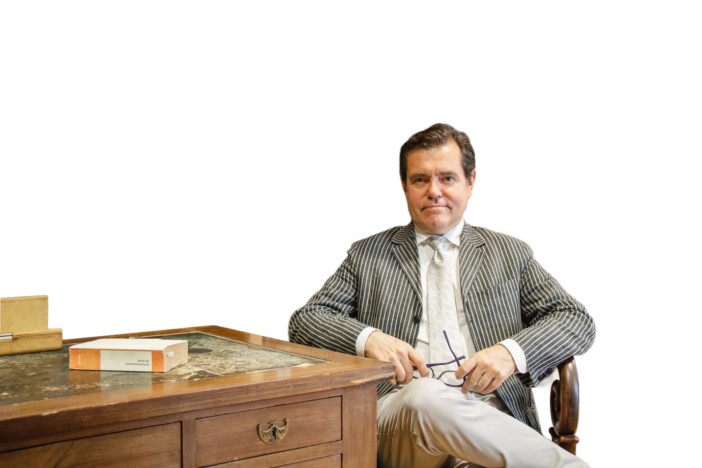 Maître Gimalac assis à un bureau en bois, portant une veste à rayures et une cravate, sur un fond blanc uni
