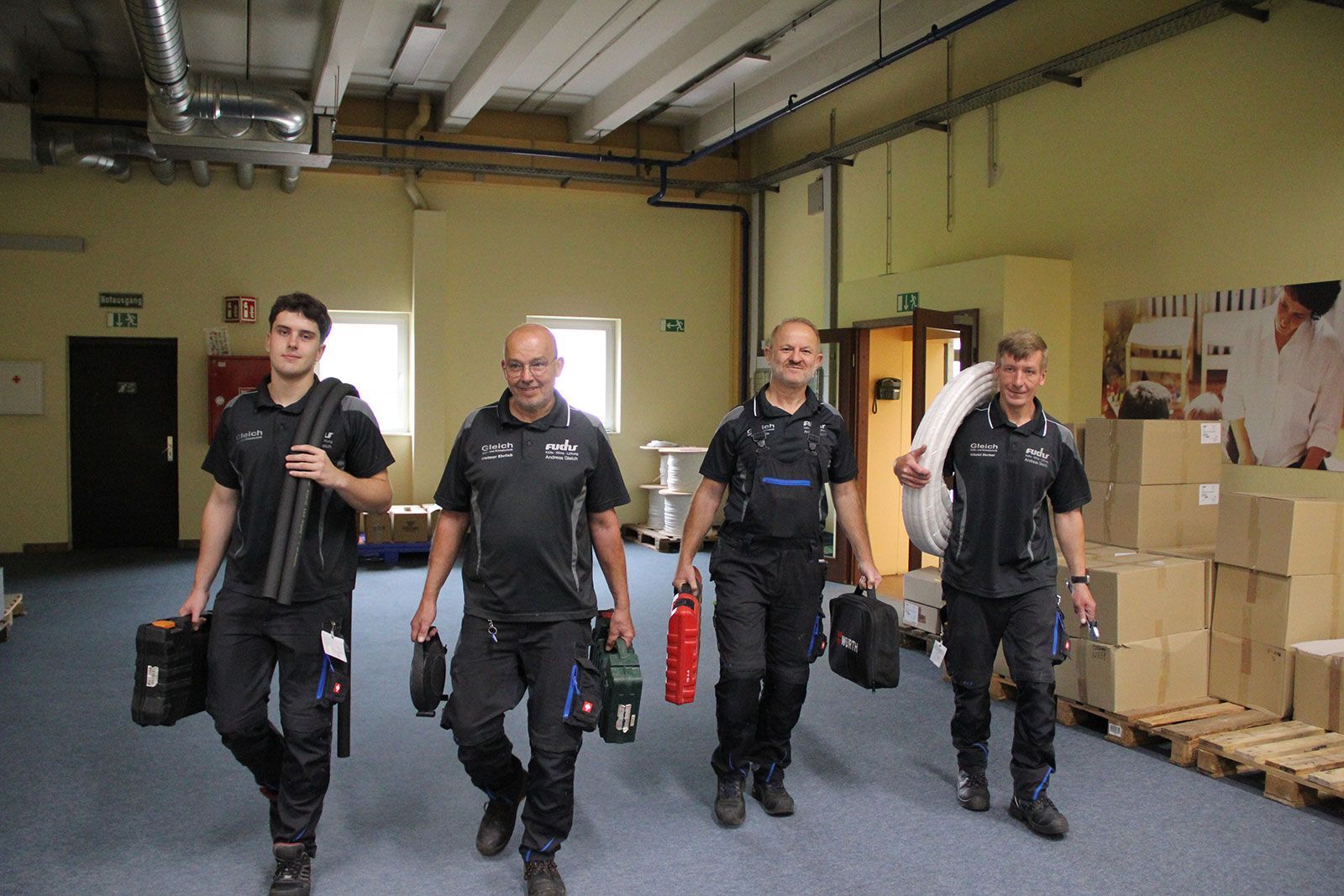 Vier Arbeiter in schwarzen Uniformen gehen mit Werkzeugen und Ausrüstung durch eine Lagerhalle.