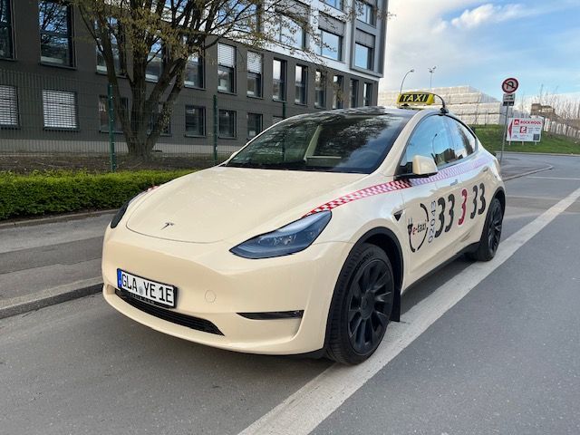 Beigefarbenes Tesla-Taxi auf einer Stadtstraße, geparkt neben Gebäuden und Bäumen.