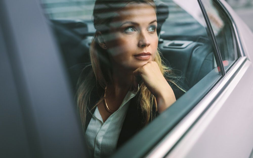 Eine Frau, die aus einem Autofenster schaut, spiegelt sich im Glas; sie trägt eine dunkle Jacke und ein weißes Hemd.