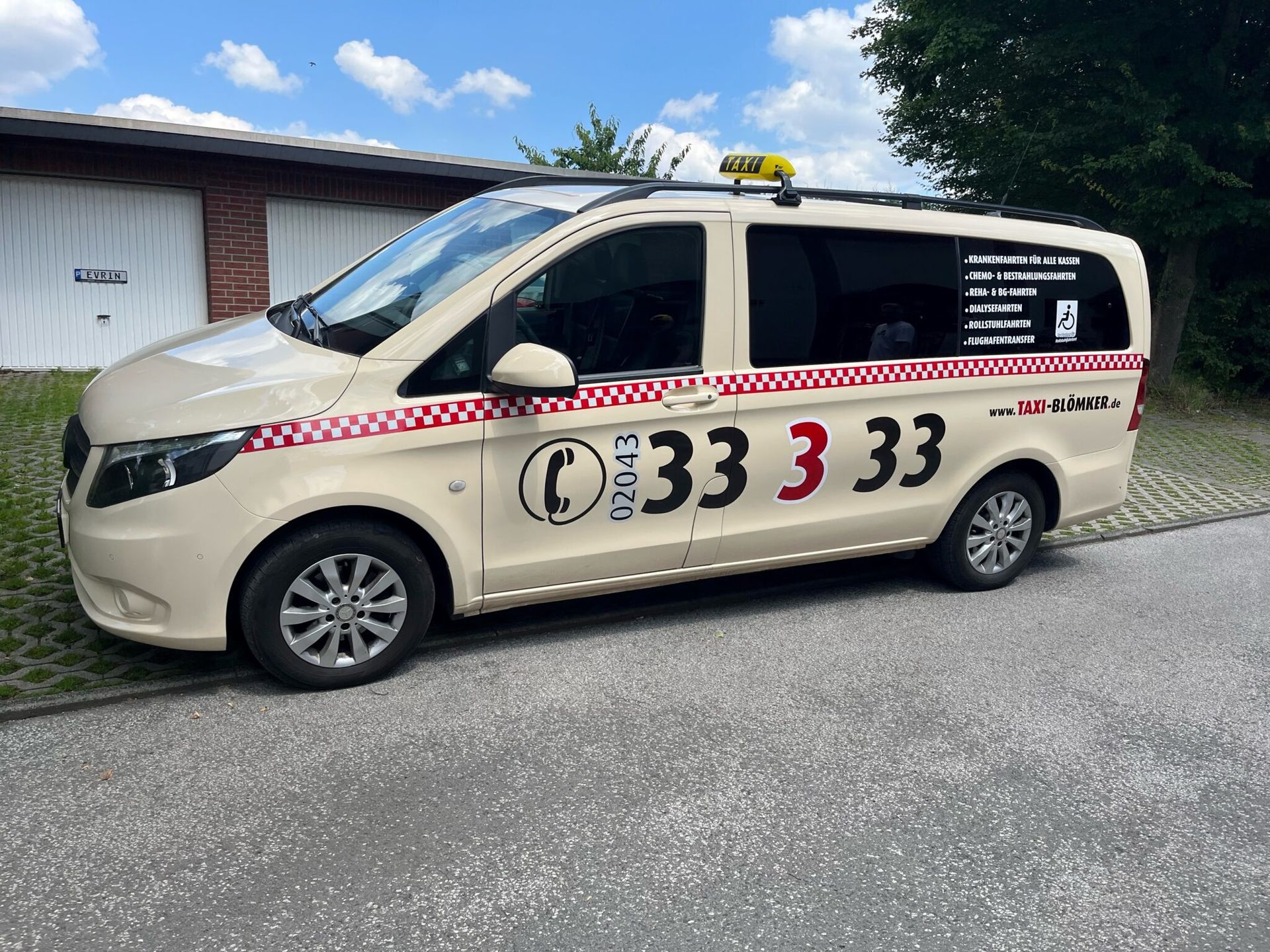 Beigefarbener Kleinbus mit rot-weiß kariertem Streifen und Telefonnummer, geparkt am Straßenrand in der Nähe eines Gebäudes.