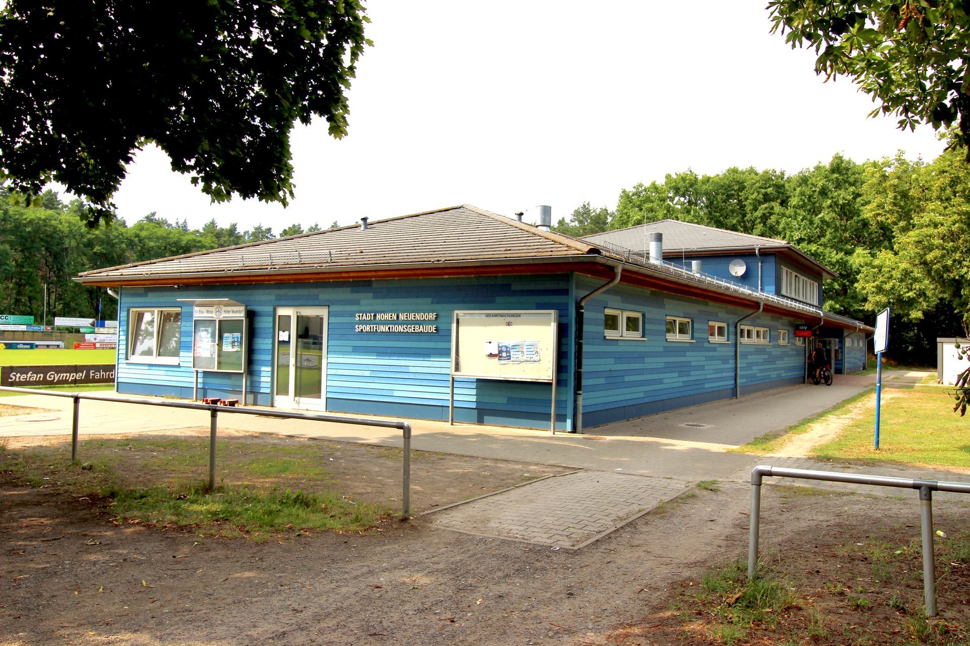 Eingangsansicht eines Sportfunktionsgebäudes mit blauer Holzfassade und umliegenden Bäumen.