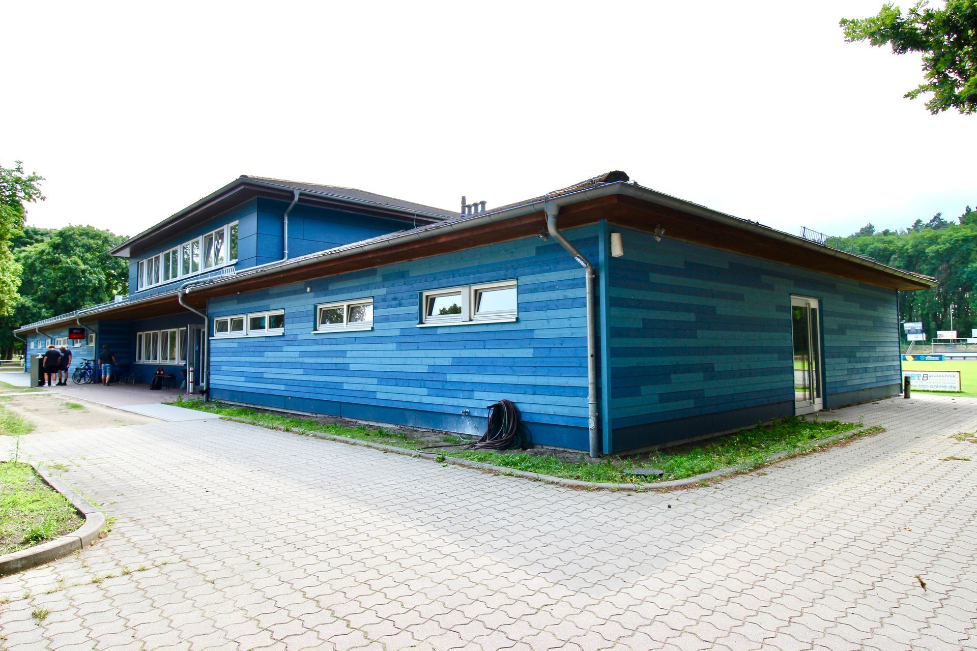 Modernes Sportfunktionsgebäude mit blauer Holzverkleidung und gepflastertem Eingangsbereich.
