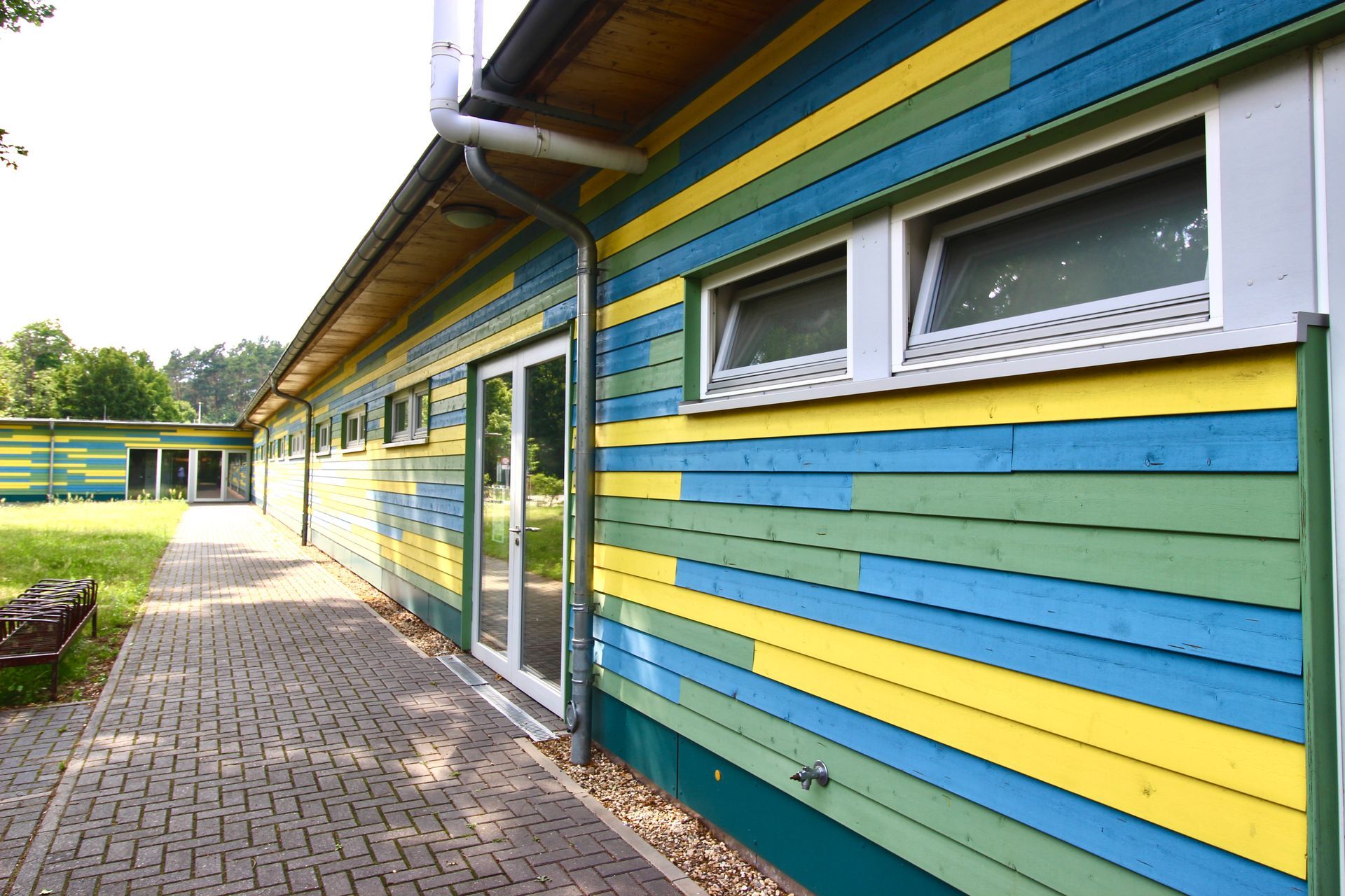 Sportfunktionsgebäude mit blau-gelber Holzfassade, Fensterband und barrierefreiem Zugang.