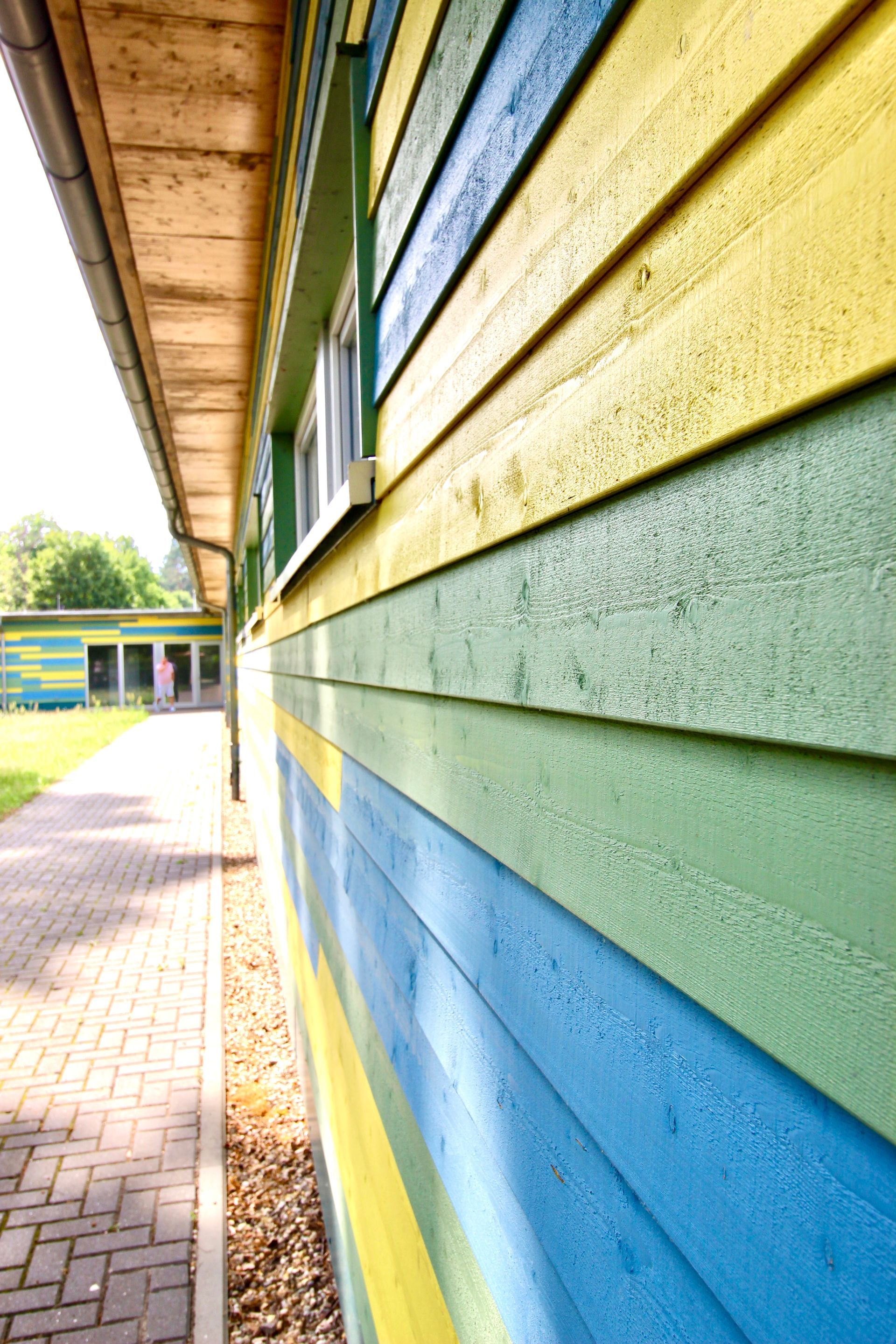 Detailaufnahme der mehrfarbigen Holzfassade mit sichtbarer Holzstruktur und Dachüberstand.