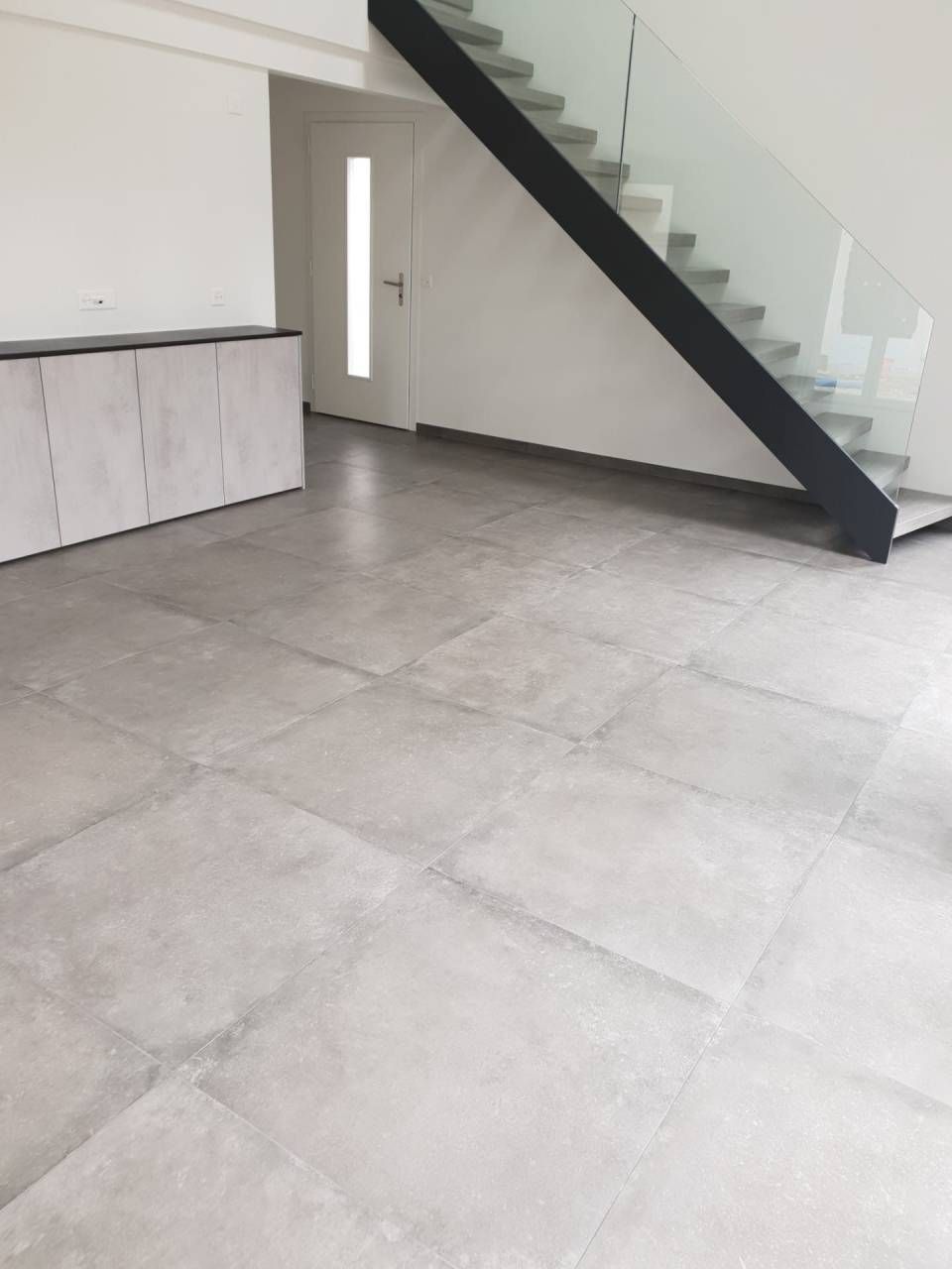 Pièce moderne et vide avec sol carrelé gris, placard intégré, escalier et porte blanche.