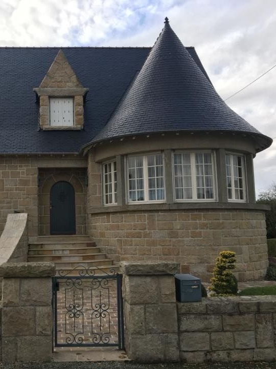 Maison en pierre avec un toit en ardoise, une tourelle avec des fenêtres et une entrée fermée par un portail.