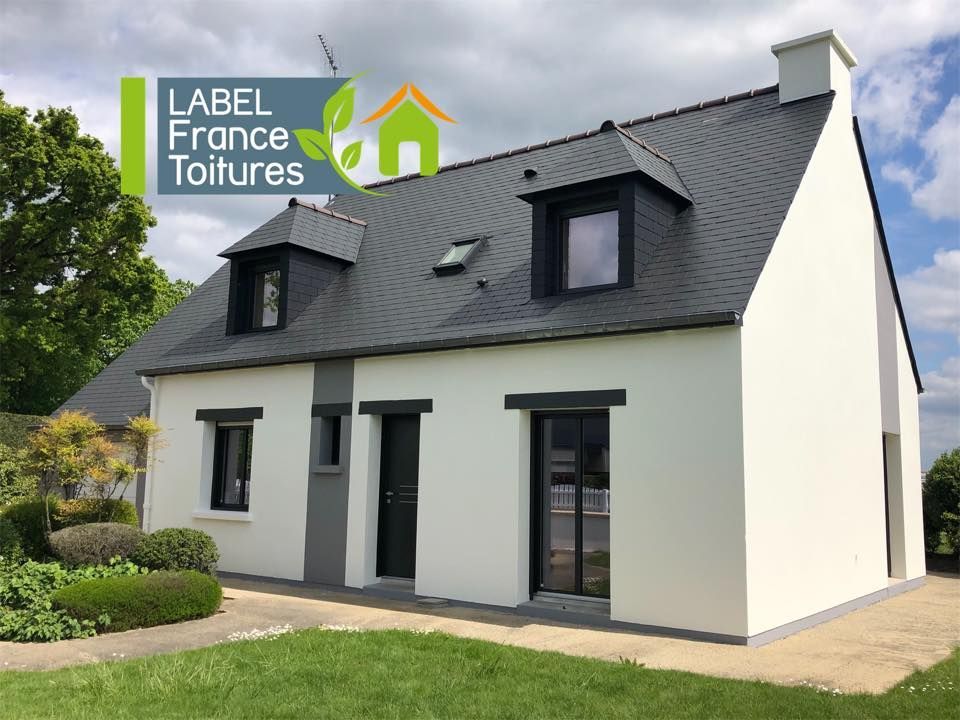 Maison beige à deux étages avec un toit en ardoise, des lucarnes et un petit auvent au-dessus de la porte d'entrée.