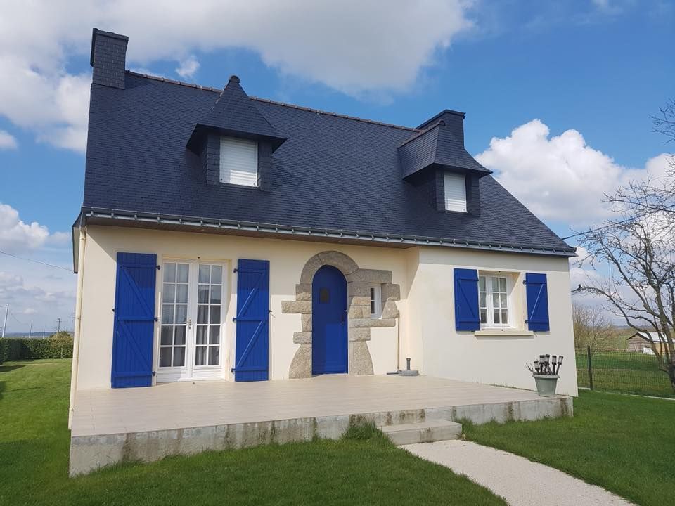 Une maison claire avec une porte bleue, des volets et un toit en ardoise, sous un ciel bleu.