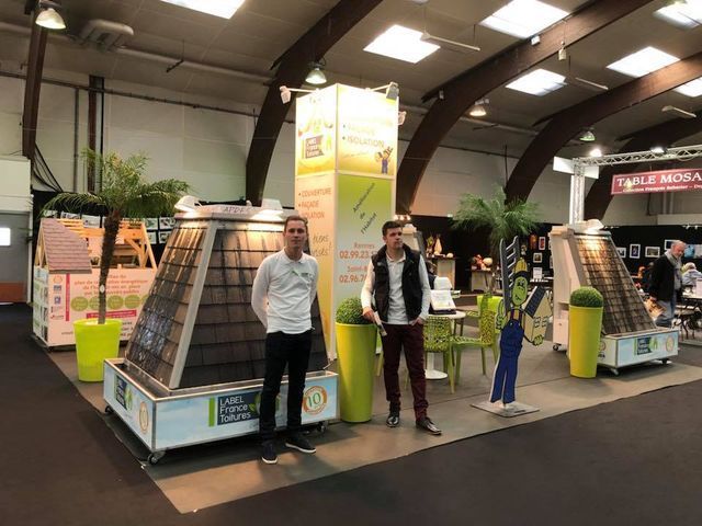 Deux hommes se tiennent près d'un présentoir de panneaux solaires entourés de végétation et de panneaux publicitaires lors d'un salon professionnel.