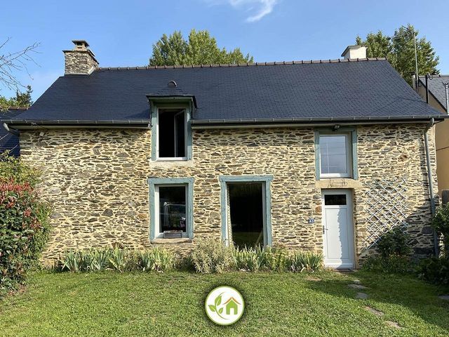 Maison en pierre au toit noir, aux fenêtres à encadrement bleu et à la porte blanche, située dans une cour herbeuse.