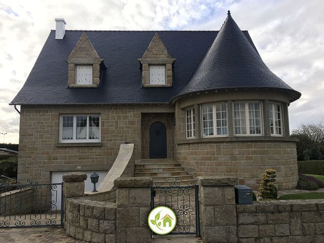 Maison en pierre avec un toit en ardoise, une tourelle et une porte cintrée derrière un mur et un portail en pierre.