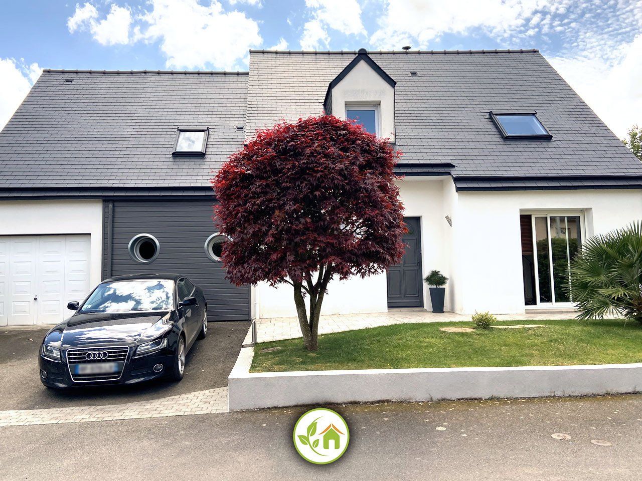 Maison moderne au toit gris, voiture noire garée devant et arbre rouge éclatant dans le jardin.