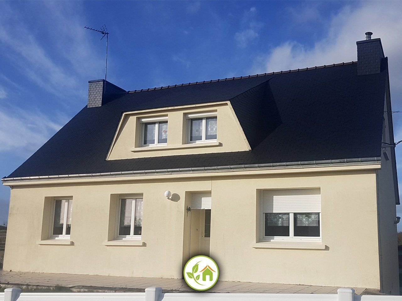 Maison beige avec un toit noir et des lucarnes sur fond de ciel bleu.