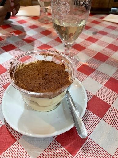 Una taza de tiramisú está en un platillo sobre una mesa al lado de una copa de vino.