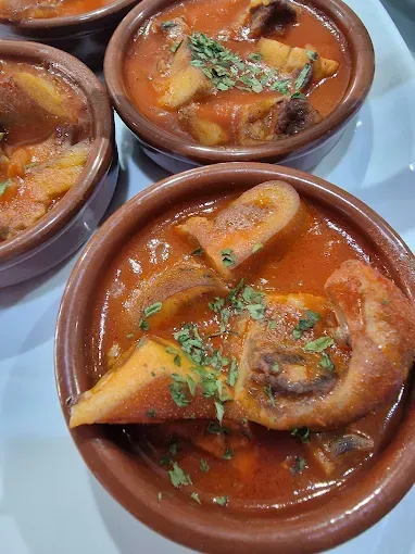 Pequeños cuencos de terracota llenos de guiso de callos en salsa roja, adornado con perejil.