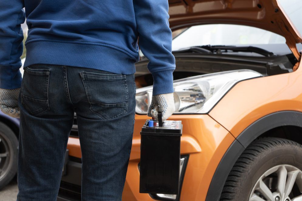 Persona sosteniendo una batería de automóvil cerca de un automóvil naranja con el capó abierto.