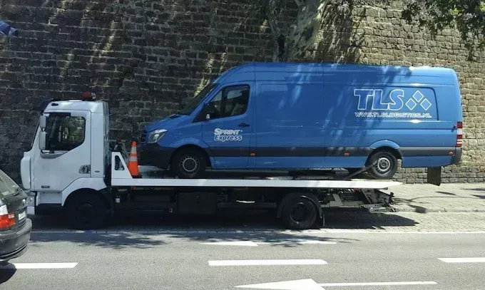 Furgoneta azul sobre una grúa de plataforma estacionada en una calle con una pared de ladrillos al fondo.