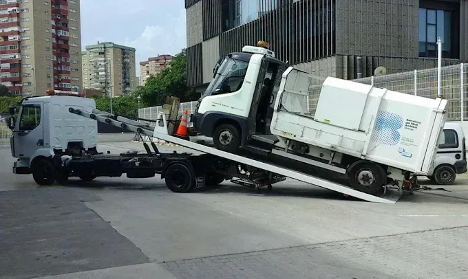 Una grúa arrastra un pequeño camión blanco por su rampa en una calle de la ciudad.