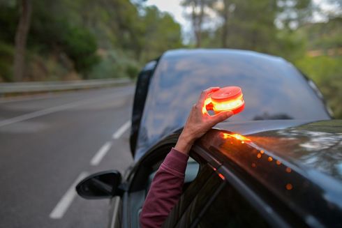 Persona sosteniendo una luz de advertencia de peligro encendida sobre un automóvil con el capó abierto.