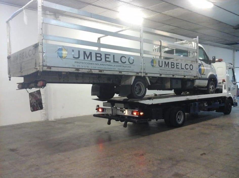 Una grúa de plataforma transporta un camión más grande. Ambos vehículos llevan el logotipo de