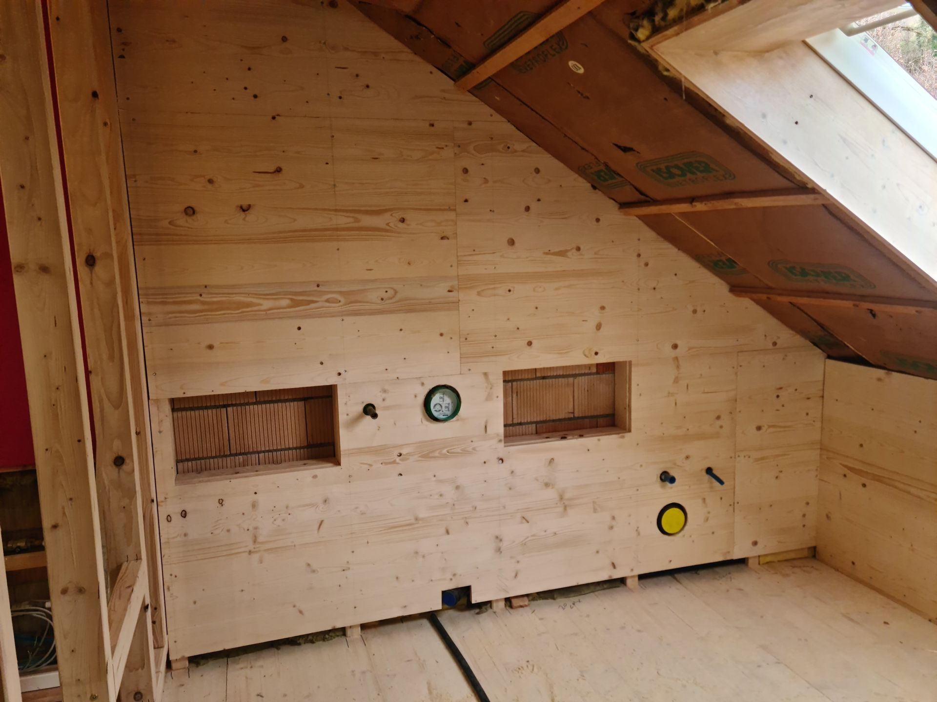 Holzgetäfeltes Dachzimmer mit Dachschräge, zwei Wandöffnungen und freiliegenden Installationsarmaturen.