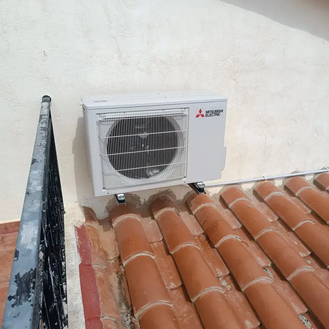 Una unidad de aire acondicionado exterior montada en una pared de color claro, sobre un techo de tejas de terracota.