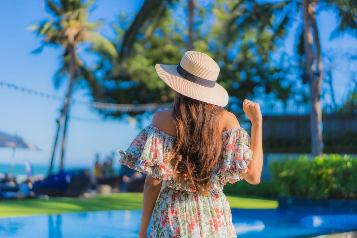 Mujer con vestido floral y sombrero mira una piscina y el océano en un día soleado.