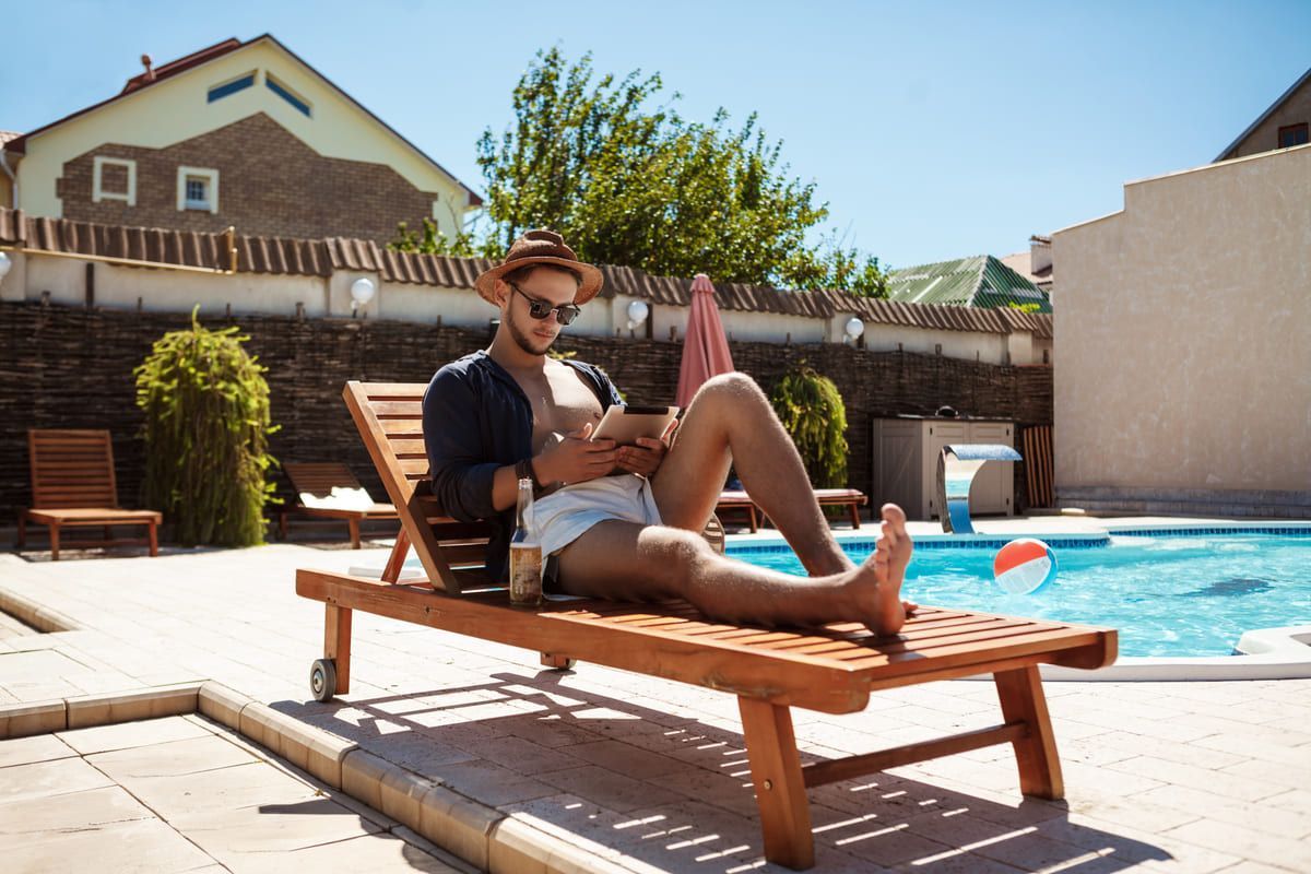 Hombre con sombrero y gafas de sol relajándose en una silla de piscina de madera, mirando una tableta cerca de una piscina.