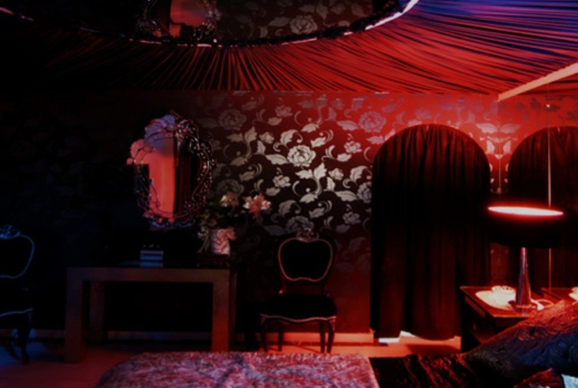 Habitación oscura y ornamentada con iluminación roja y azul. Incluye papel tapiz floral, un espejo y una puerta arqueada.