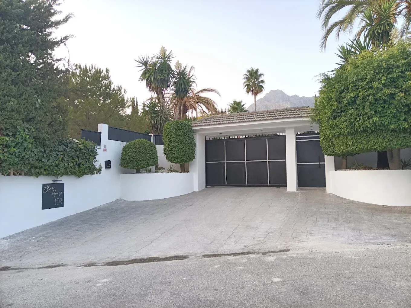 Entrada con portón negro a una propiedad de paredes blancas con camino de entrada y árboles.