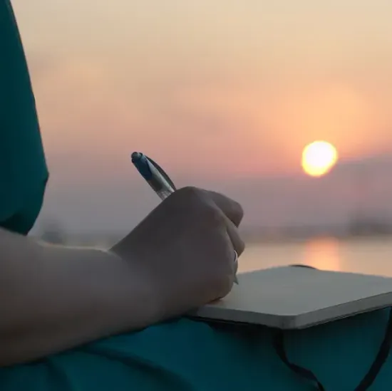 Una persona está escribiendo en un cuaderno con un bolígrafo frente a una puesta de sol.
