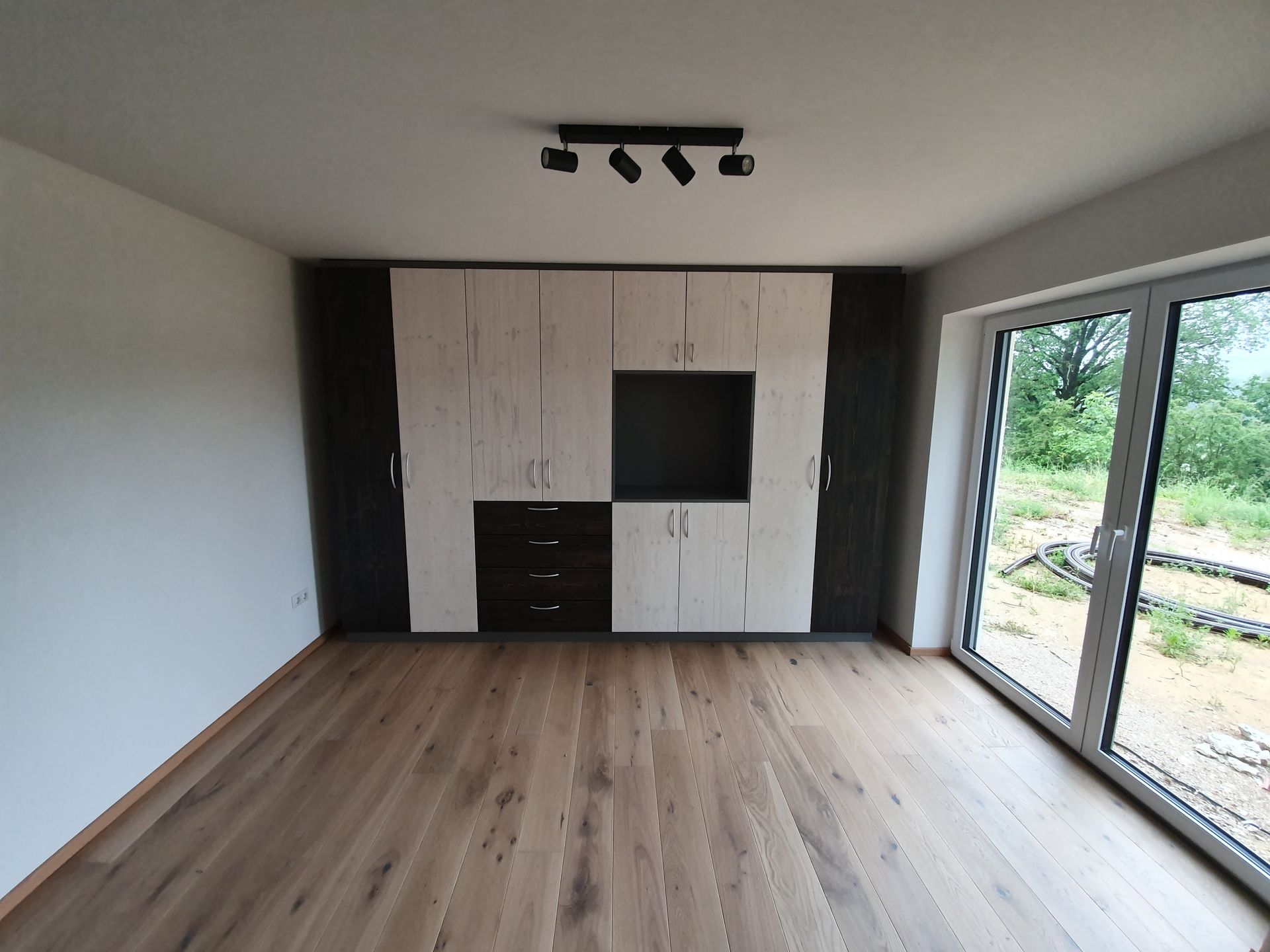 Leerer Raum mit hellem Holzboden, großem Schrank mit hellen und dunklen Paneelen, Glasschiebetür, Deckenschienenbeleuchtung.