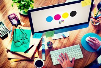 Escritorio de diseñador con un monitor que muestra círculos de colores, teclado, ratón, cuaderno y taza de café.
