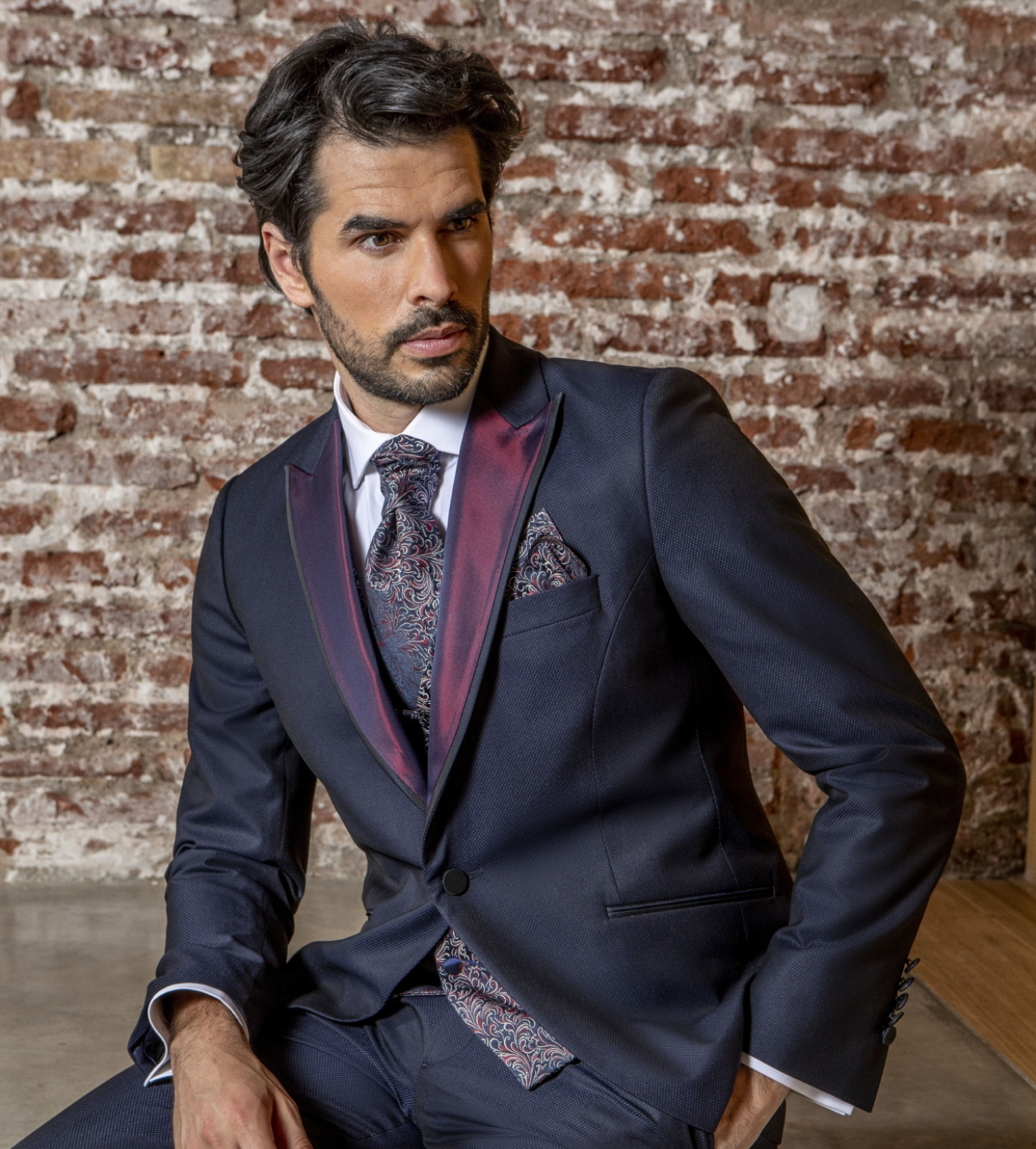 Un hombre con traje y corbata está sentado frente a una pared de ladrillos.