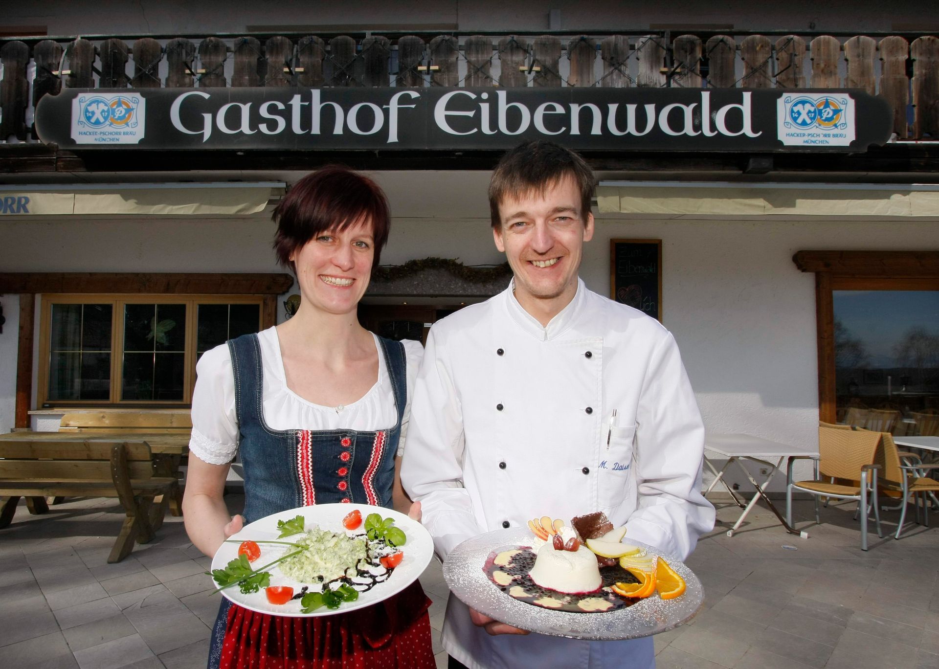 Wirtspaar in Tracht vor dem Gasthof Eibenwald mit angerichteten Speisen.