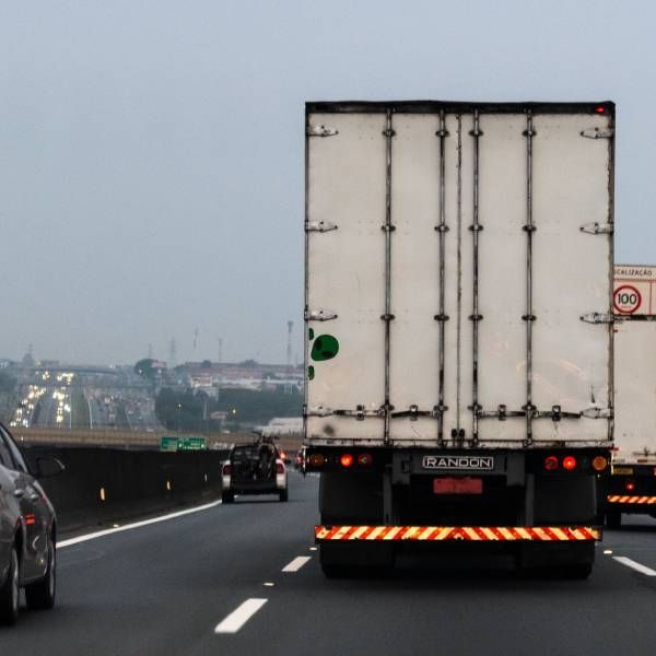 Operário descarregando mercadorias empilhadas de um caminhão branco estacionado ao lado de uma parede amarela.